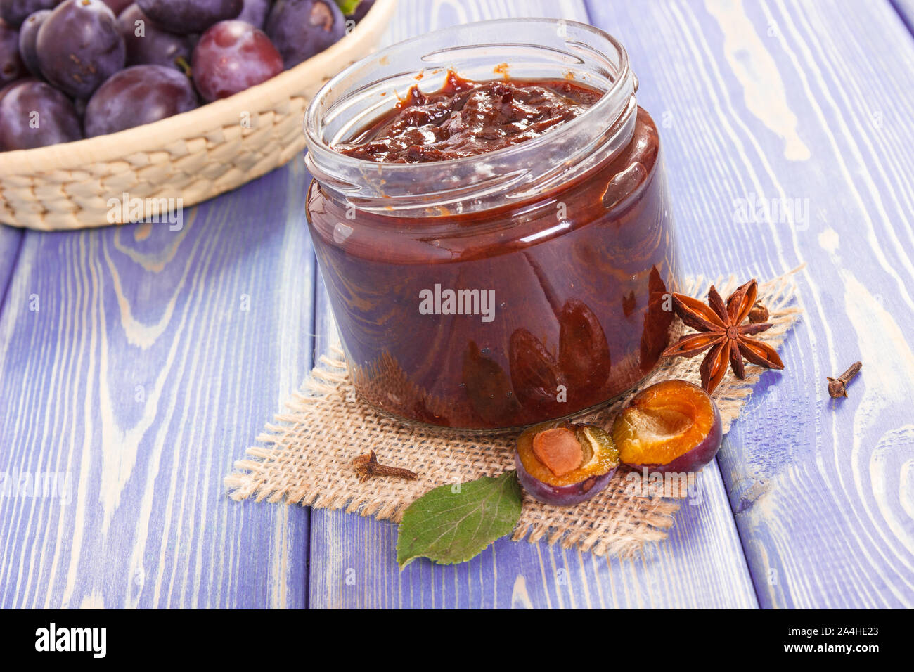 Fresh homemade plum marmalade in glass jar, spices and ripe fruits in wicker basket in background, concept of healthy sweet dessert Stock Photo