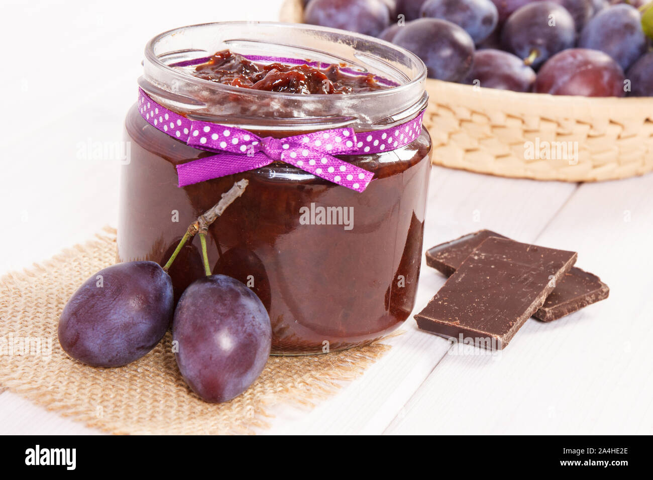 Fresh plum jam or marmalade in glass jar, ripe fruits in wicker basket and chocolate, concept of healthy sweet dessert Stock Photo