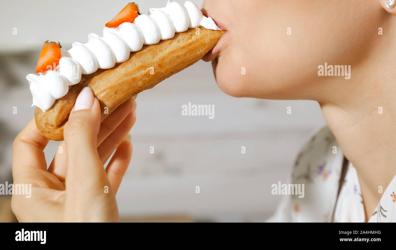 pretty young woman eats beautiful tasty eclair with cream and strawberry slices on blurred background extreme close view Stock Photo