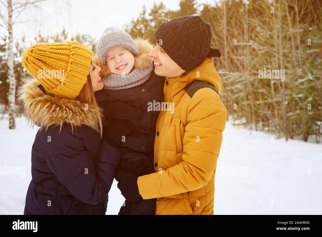 Portrait of happy family in winter day. Mom and dad are cuddling and kissing their little son in winter park. Family lovely moments, copy space Stock Photo