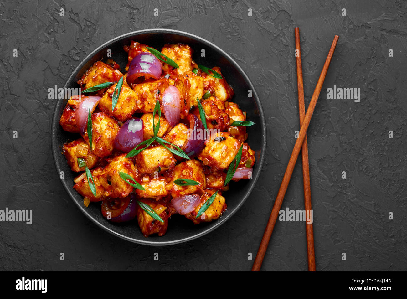 Schezwan Paneer in black bowl at dark slate background. Schezwan Paneer is indo-chinese cuisine dish with deep fried Paneer cheese, onion and Schezwan Stock Photo
