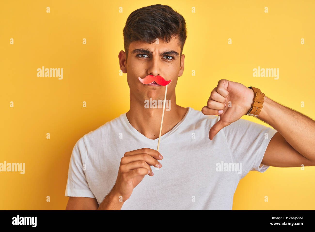 Young indian man holding fanny mustache standing over isolated yellow ...