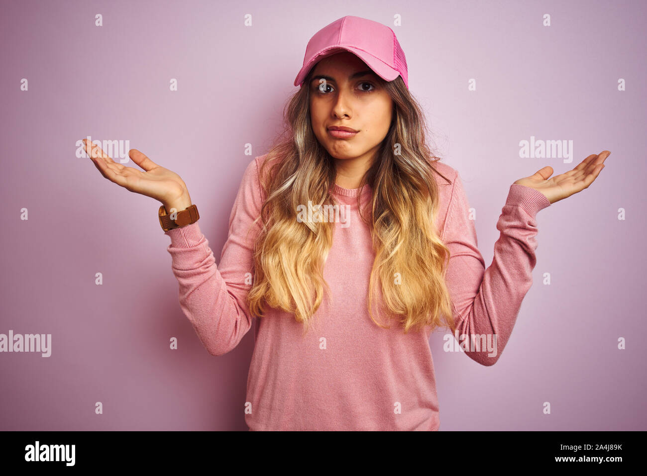 Young beautiful woman wearing cap over pink isolated background clueless and confused expression with arms and hands raised. Doubt concept. Stock Photo
