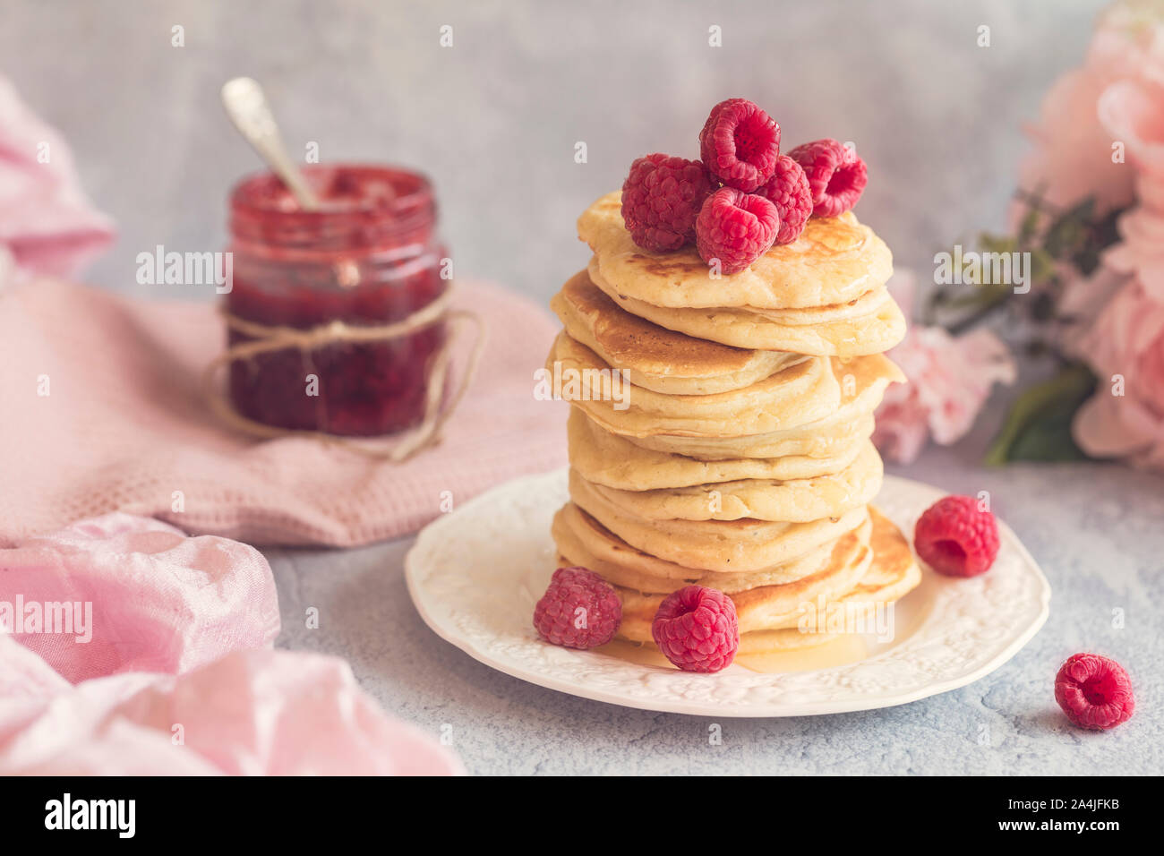 Pancake stack with fresh raspberries on top. There is a jar of jam, some flowers and a pink fabric in the background. The background is in mixed shade Stock Photo