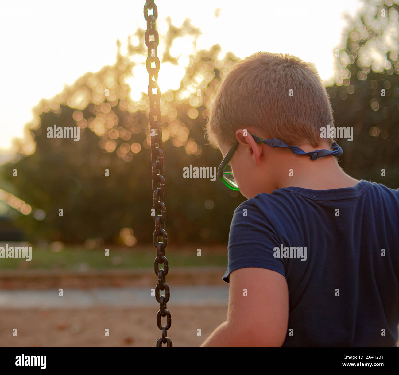 Blond boy with glasses looking at the floor while sitting on a swing in a playground Stock Photo