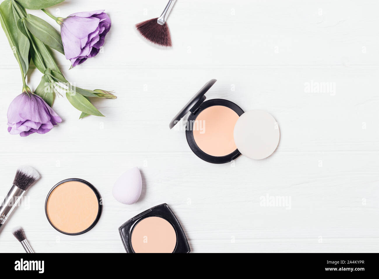 Compact powder and sponge applicator next to brushes, cosmetic products and fresh purple lisianthus on white background, flat lay composition. Stock Photo
