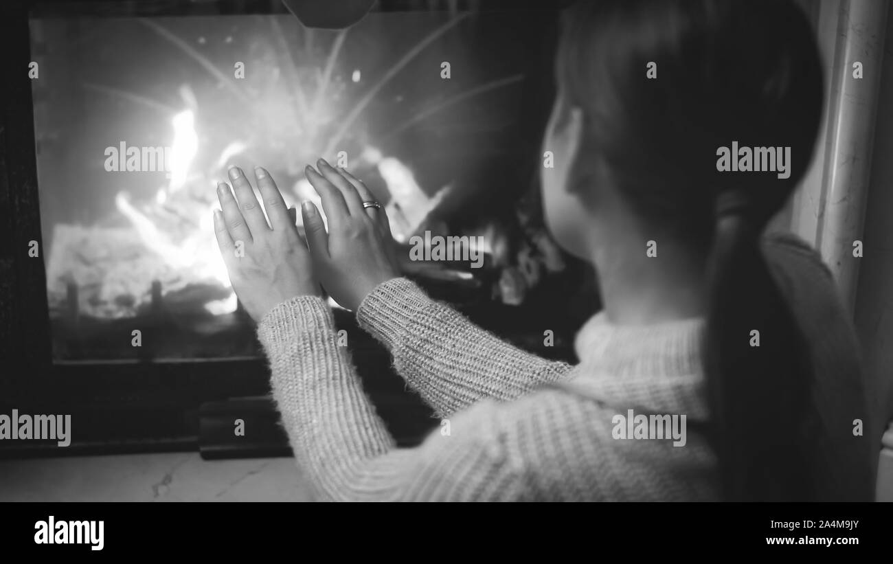 Black and white closeup image of woman sitting by the fireplace and warming her hands Stock Photo