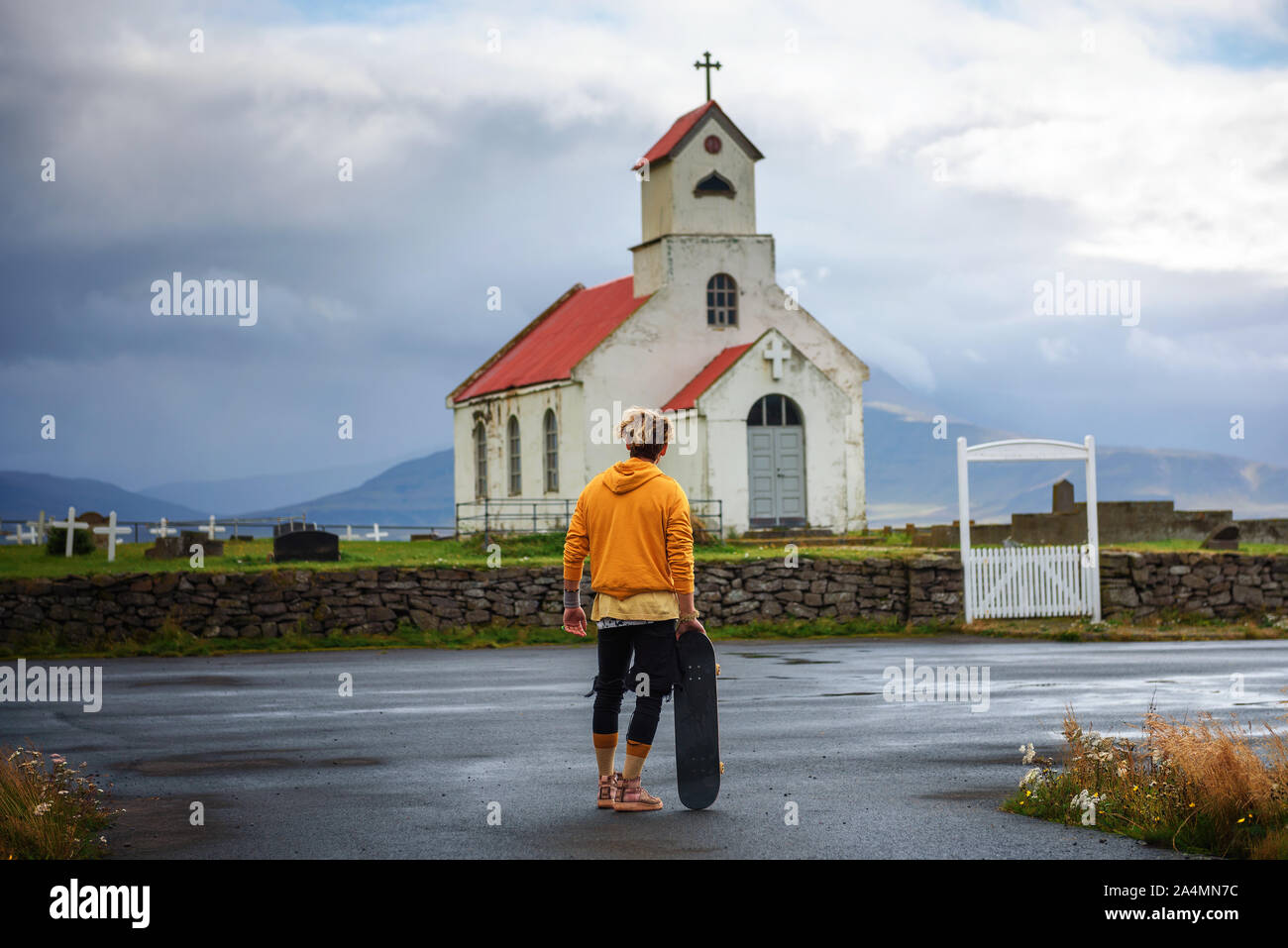 Young man with a skateboard looking at a church with a cemetery in Iceland Stock Photo
