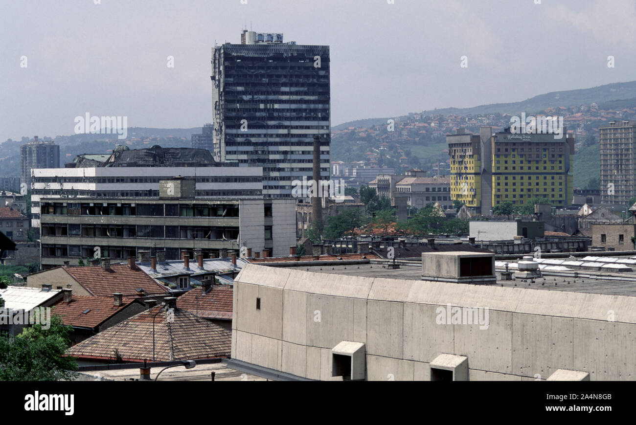 5th June 1993 During the Siege of Sarajevo: the view north-west from Stolačka Street. The burned-out tower block of the Building of the BiH Parliamentary Assembly dominates the frame with the Holiday Inn Hotel on the right. In front of the Assembly Building are the ruins of the old tobacco factory. Stock Photo