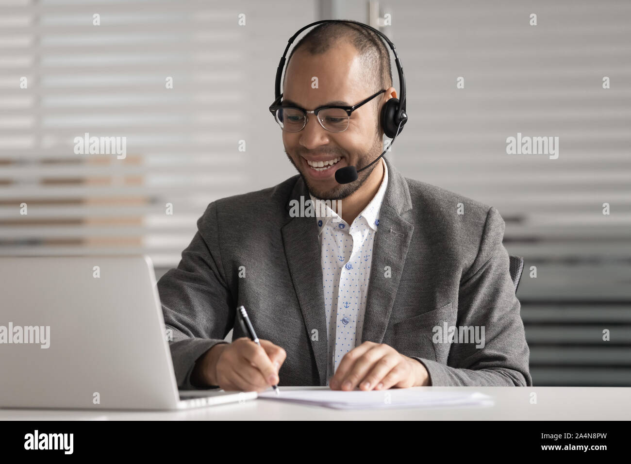 Happy African American employee in headset making notes, using laptop ...
