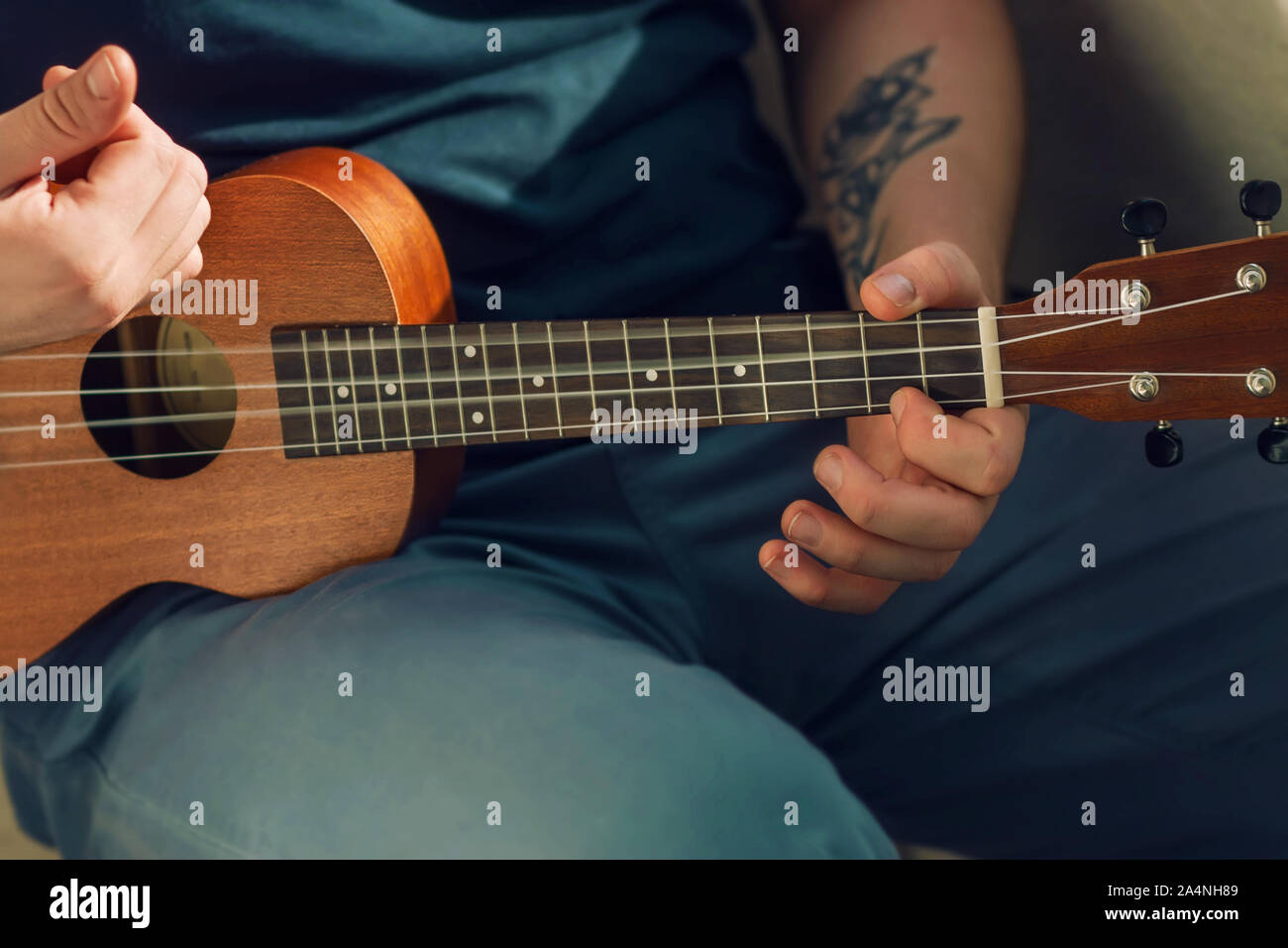A man in blue clothes and with a tattoo on his arm plays a cheerful tune on a ukulele on a Sunny day. Stock Photo