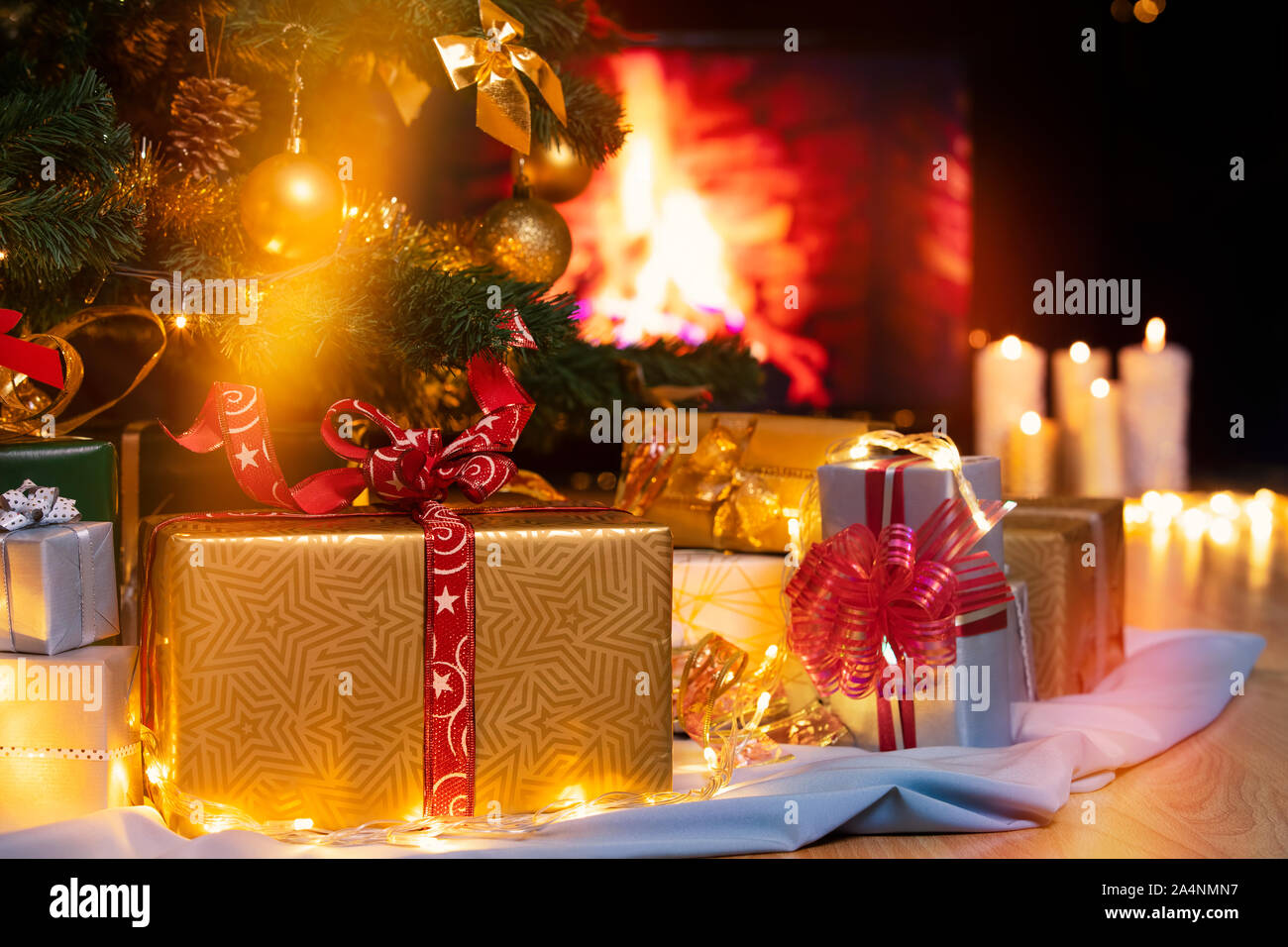Stack of packed gift boxes under Christmas tree against burning fireplace. Lots of Christmas gifts under the tree. Candles on wooden floor. Focus on g Stock Photo