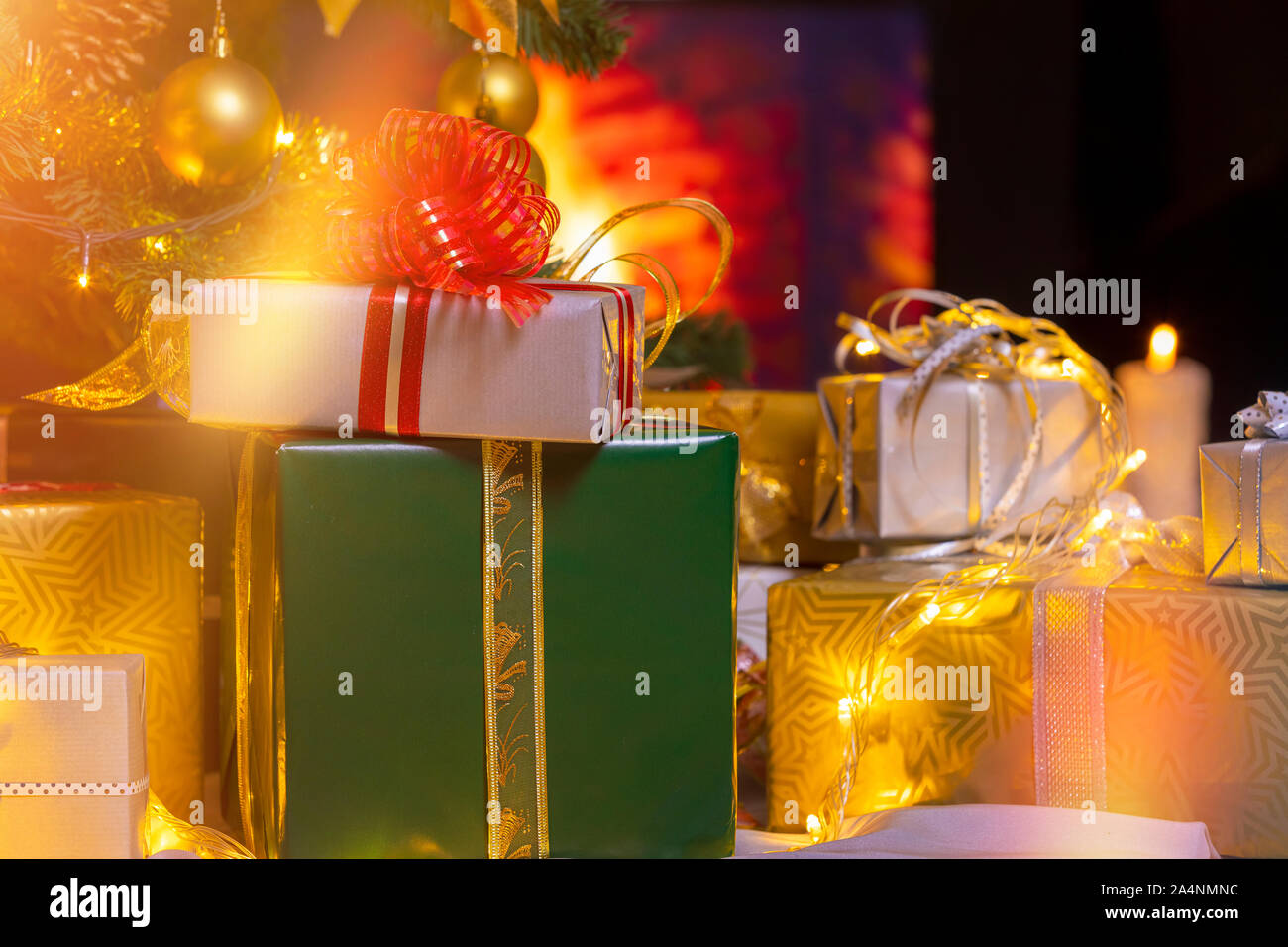 Stack of packed gift boxes under Christmas tree against burning fireplace. Lots of Christmas gifts under the tree. Candles on wooden floor. Focus on g Stock Photo