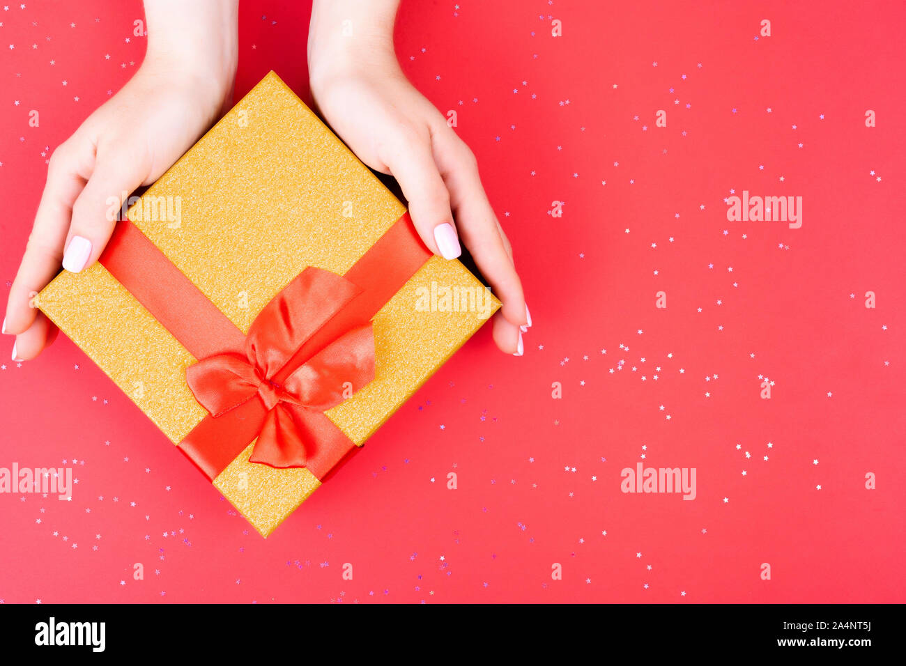 Female hands hold a gift box of gold color with a bow on a red background. View from above. Place for text. Stock Photo