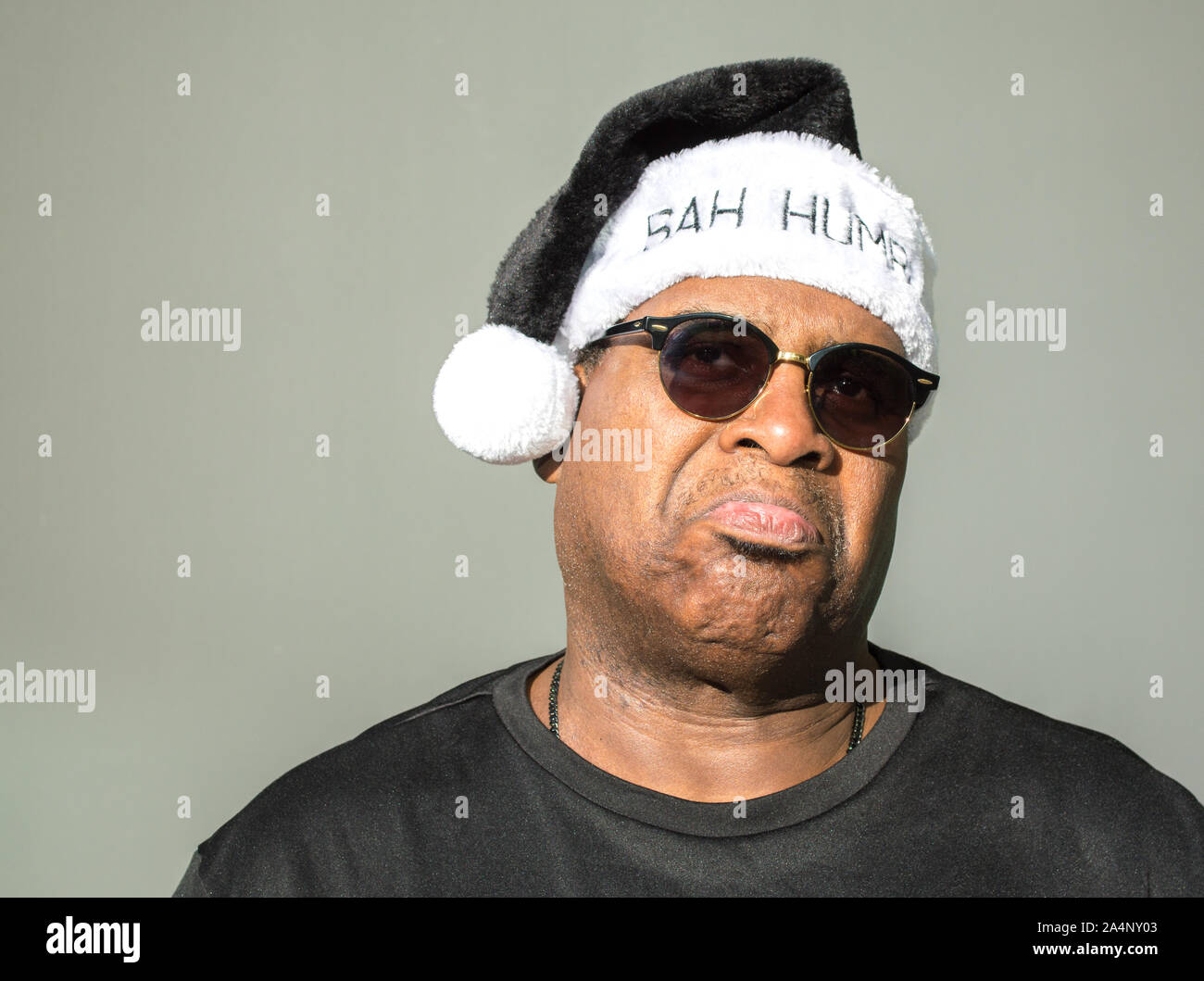 a frowning middle aged African American man wearing a black and white Santa Claus hat saying Bah Humbug on it against a solid background Stock Photo