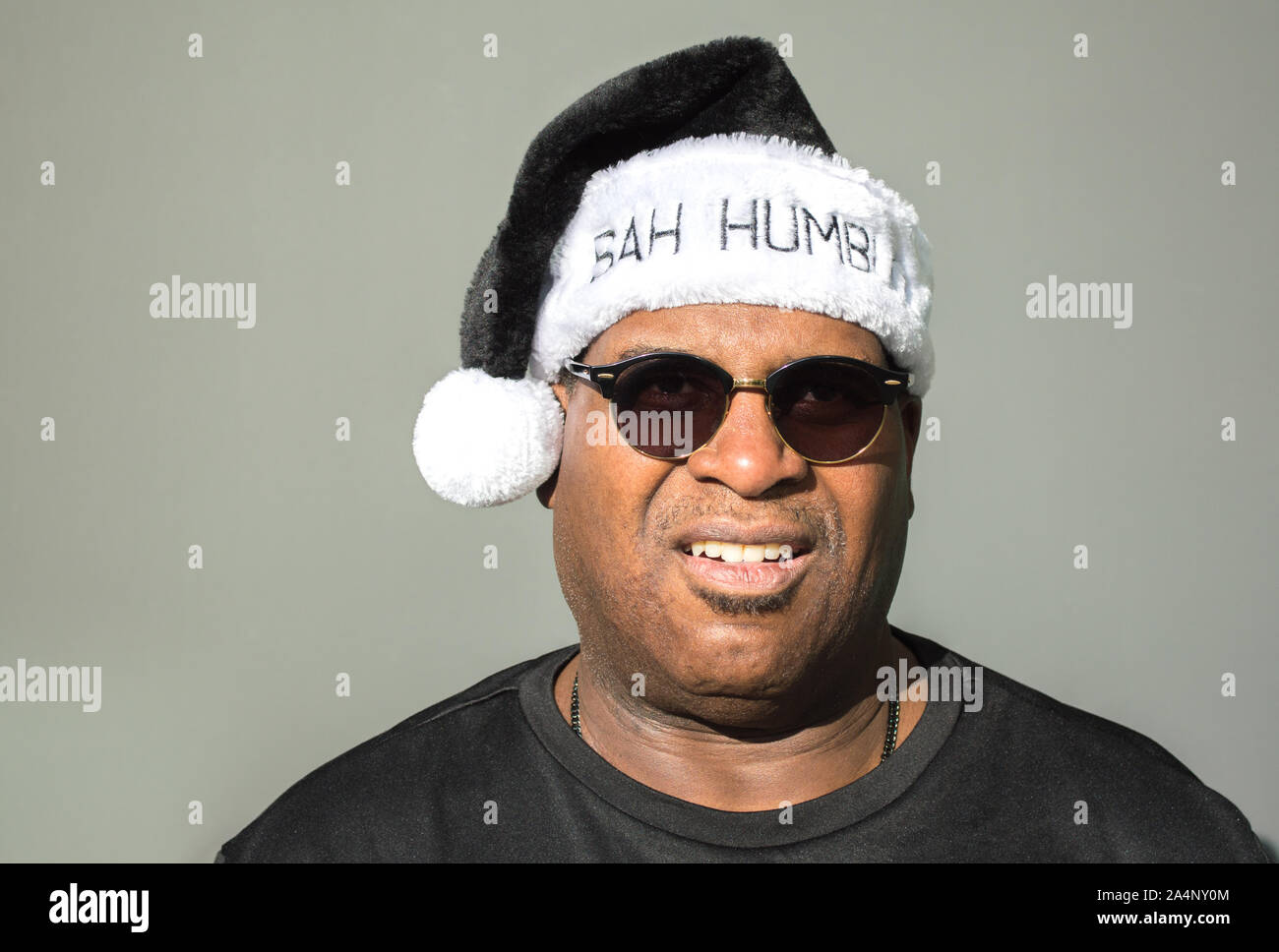 a frowning middle aged African American man wearing a black and white Santa Claus hat saying Bah Humbug on it against a solid background Stock Photo