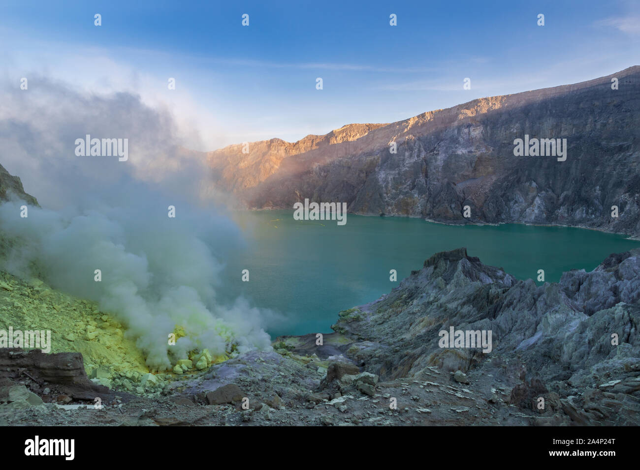 Acid lake in the crater of Mount Ijen, East Java, Indonesia. Smoke and sulfur deposits on shore from volcanic vents. Blue sky behind far crater wall. Stock Photo