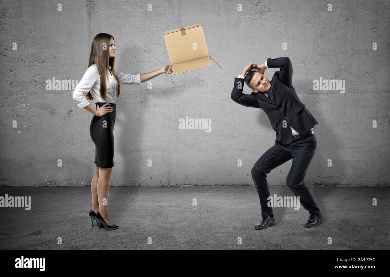 Businesswoman on concrete background holding a carton box to a cowering man. Stock Photo