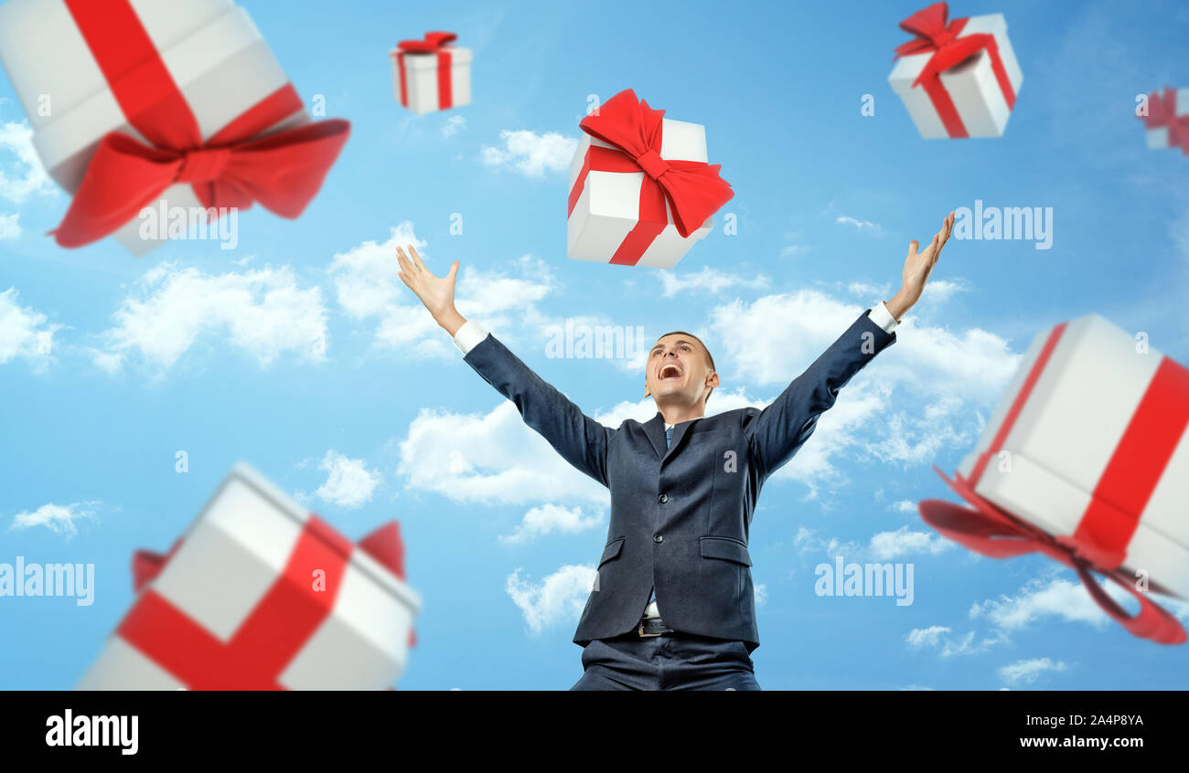 A happy businessman standing with hands raised in victory motion under a rain of gift boxes falling on him. Stock Photo