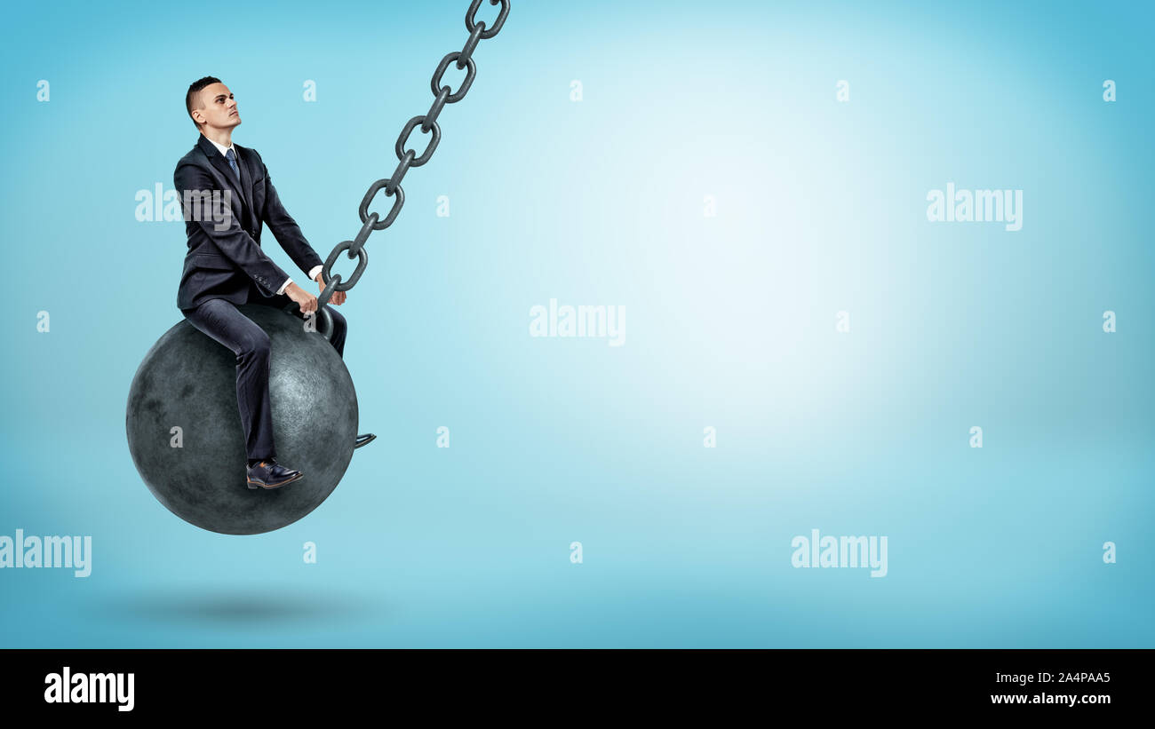 A businessman swinging on a large wrecking ball and looking up on blue background. Stock Photo