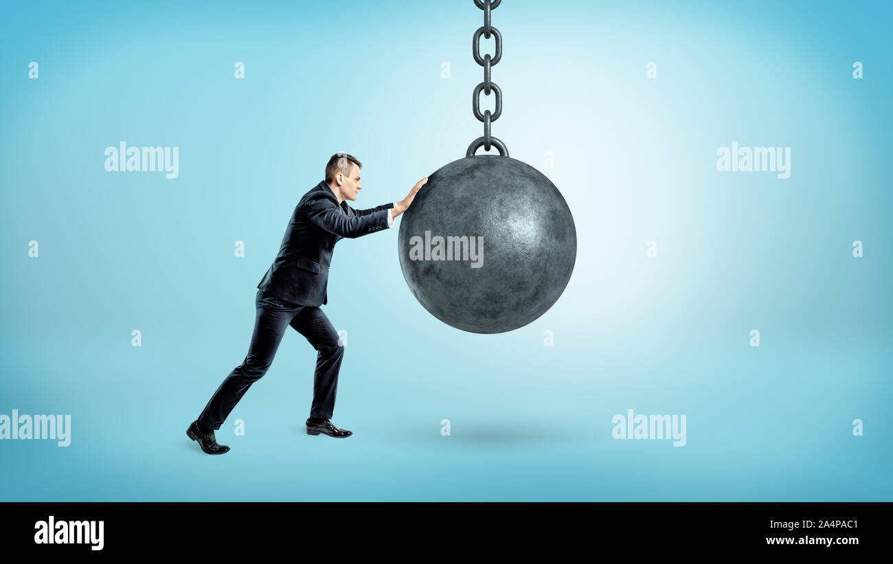 A businessman on blue background pushing at a hanging unmoving wrecking ball. Stock Photo
