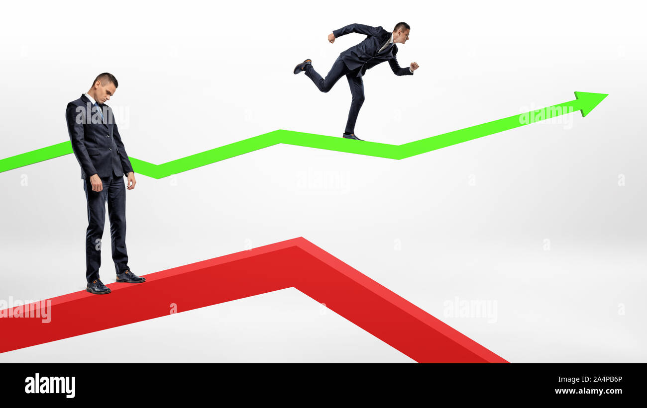 A sad businessman stands on a red decreasing arrow while another man runs up a green upwards arrow. Stock Photo