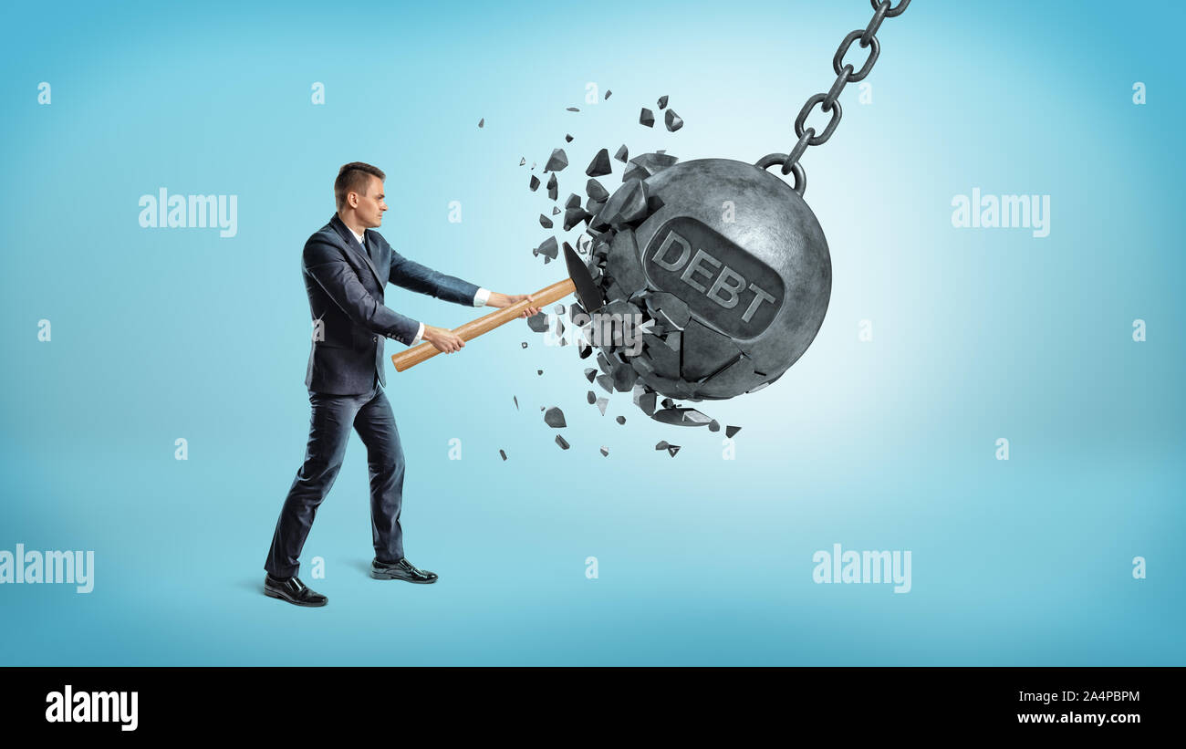 A small businessman smashes a giant swinging iron ball with a word DEBT on it using a hammer. Stock Photo