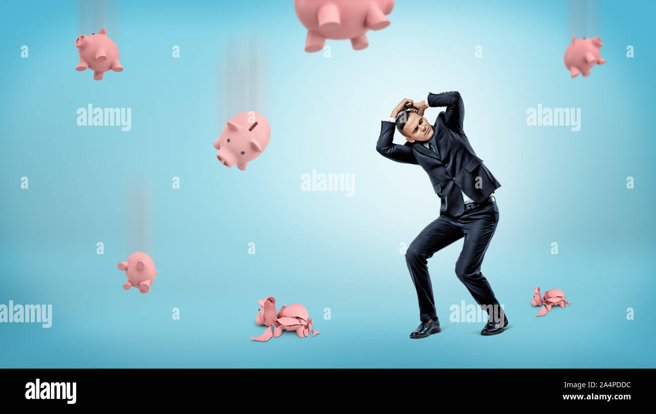 A businessman hides his head from many piggy banks that fall down and crack at the floor. Stock Photo