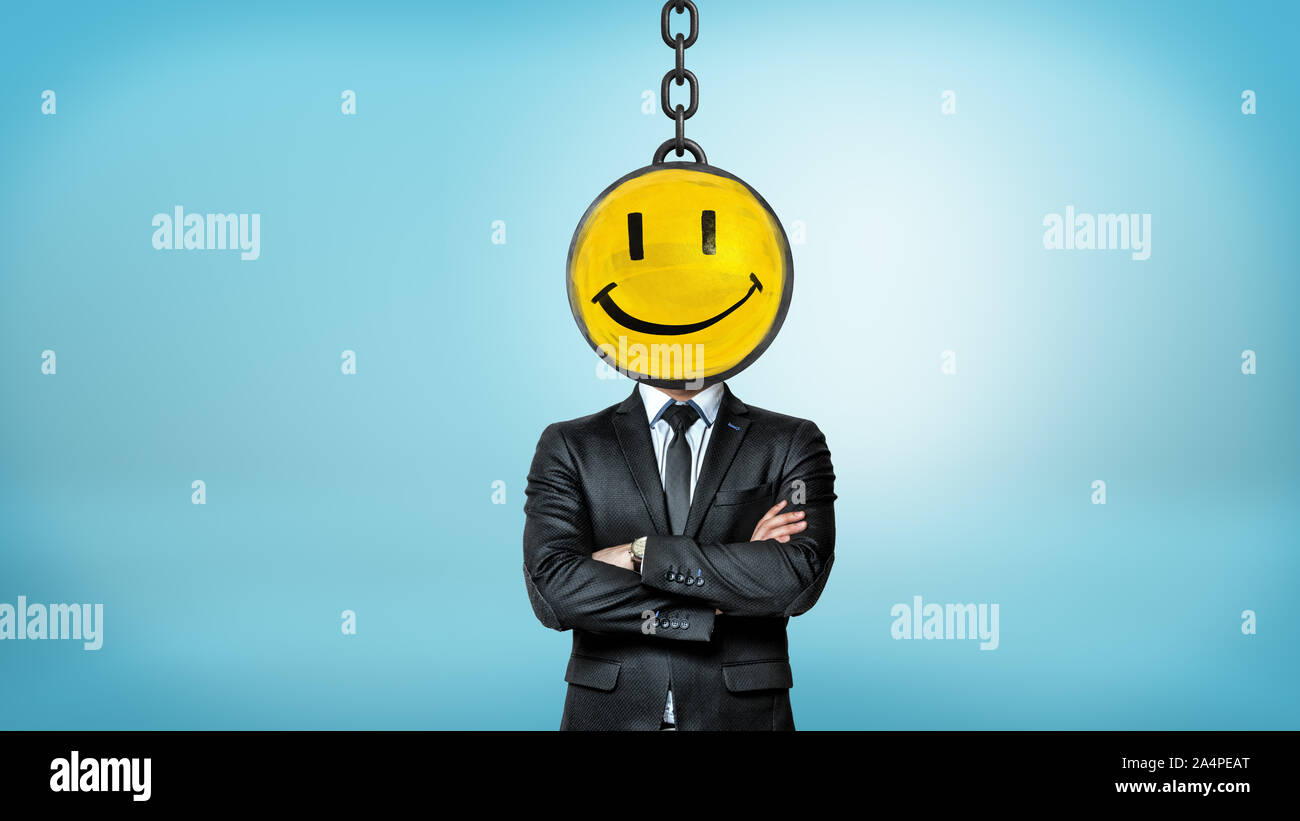 A businessman stands with folded arms with a head hidden by a wrecking ball painted with a happy face. Stock Photo