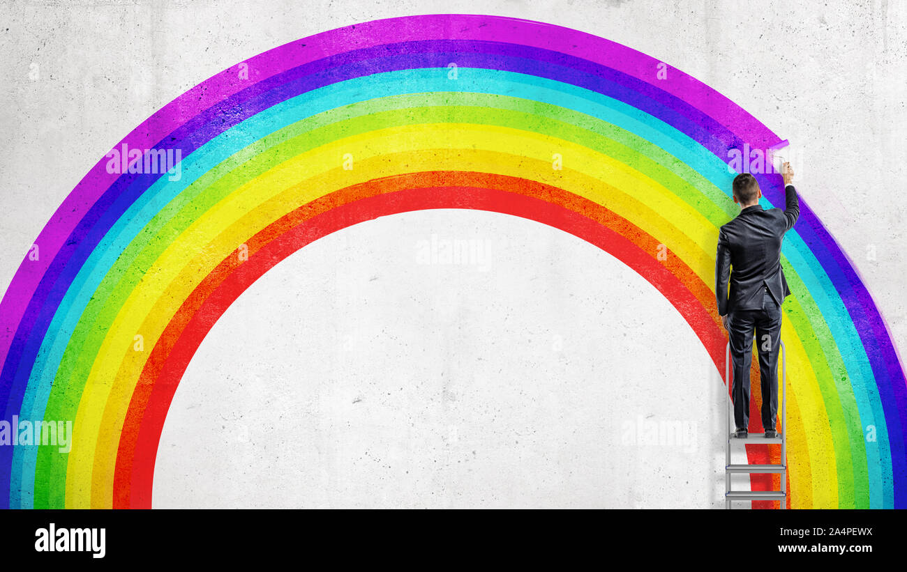 A businessman stands on a stepladder and drawing the last colorful line in the wall picture of a rainbow. Stock Photo