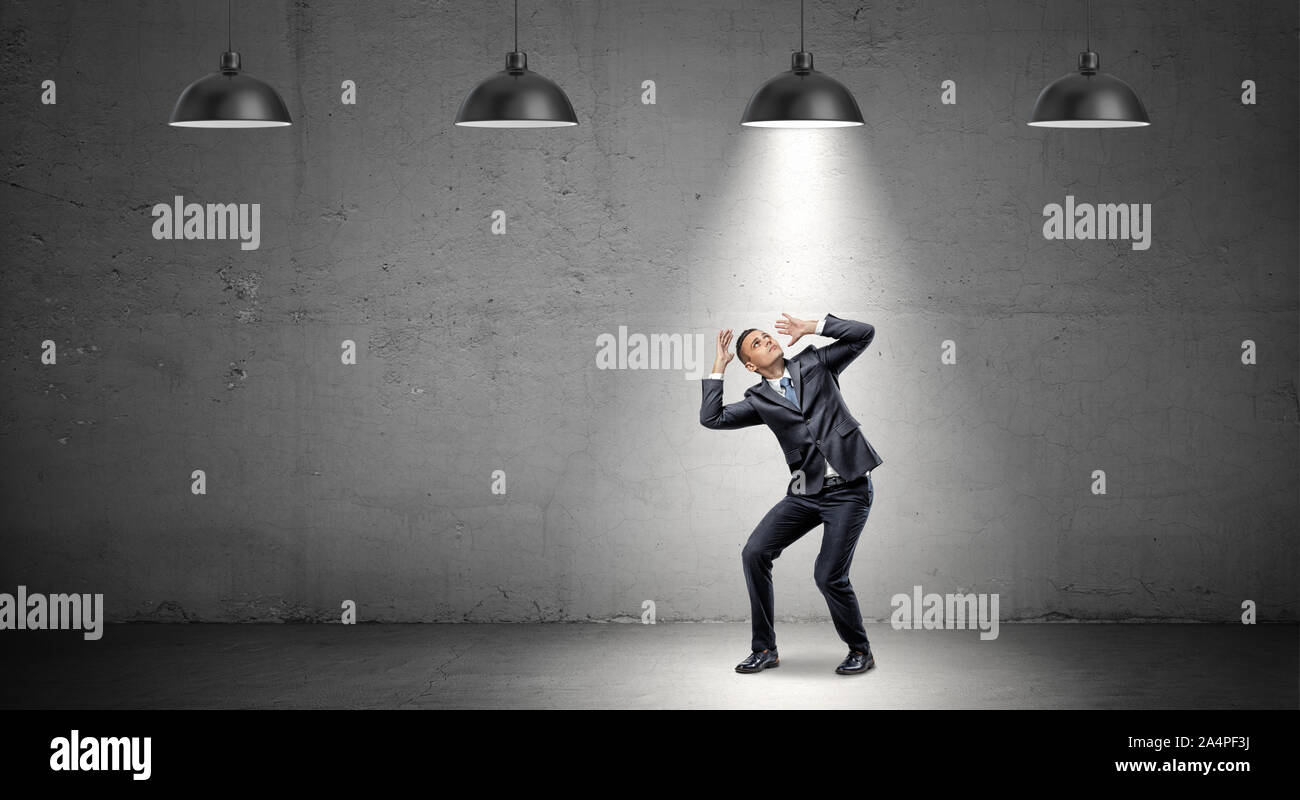 A scared businessman stands cowering under the rays of industrial pendant lights right above him. Stock Photo