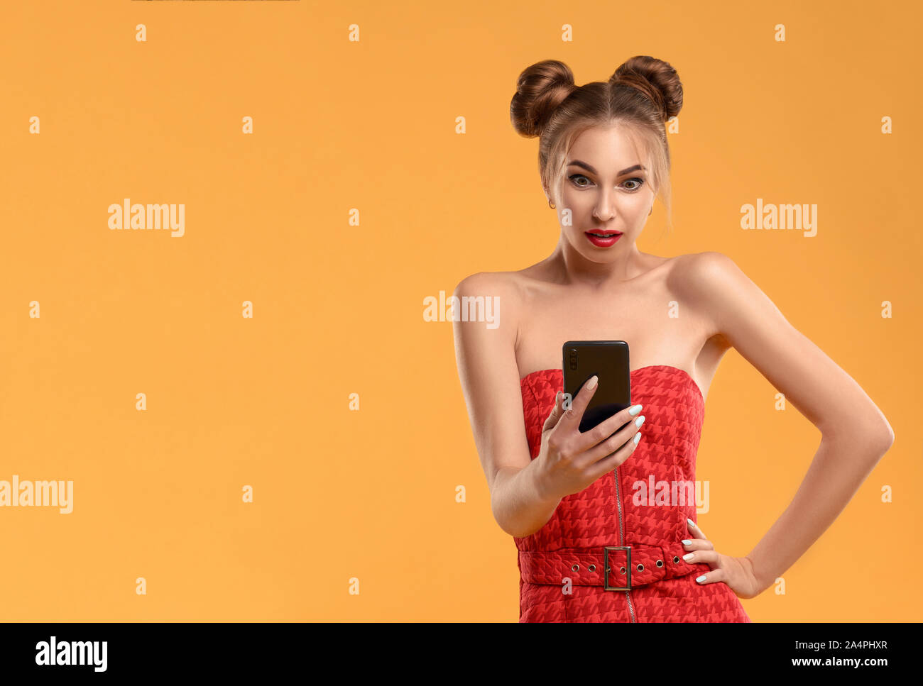 Girl with hair buns holding a smartphone with surprised face isolated on orange background Stock Photo
