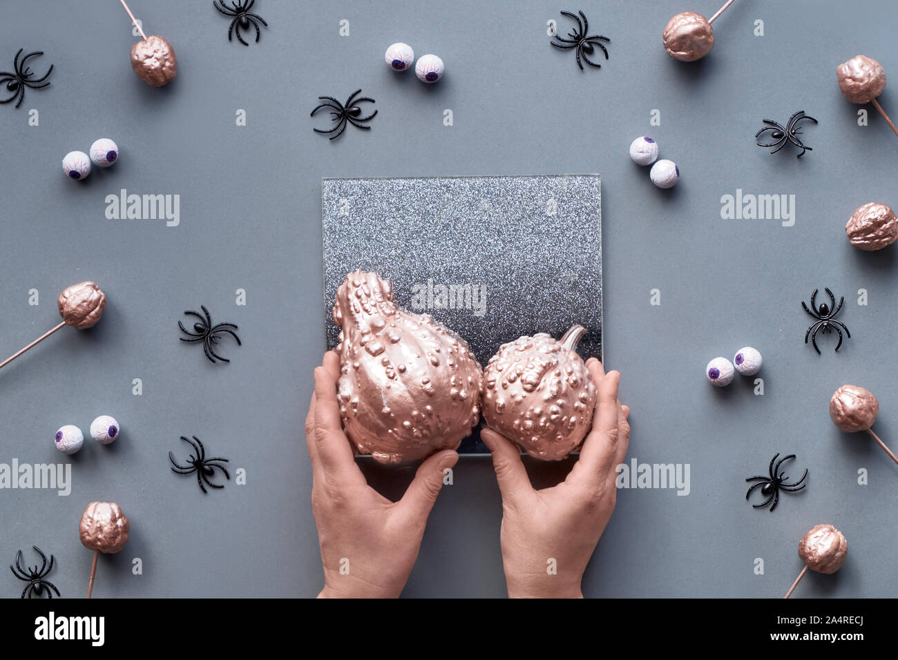 Hands trying to fit two pumpkins on decorative mirror. Creative Halloween flat lay background in grey, pink and black with chocolate eyes and spiders. Stock Photo