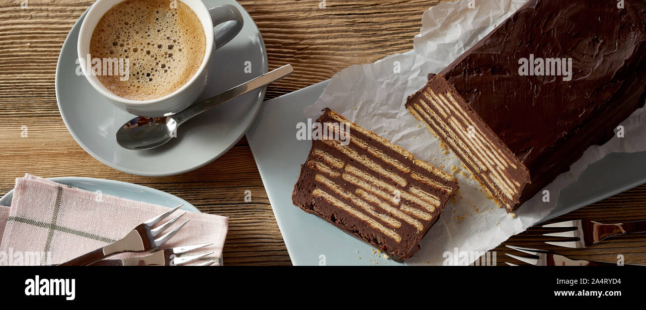 Overhead view of chocolate iced biscuit cake with one slice cut and a blue ceramic cup of creamy coffee Stock Photo