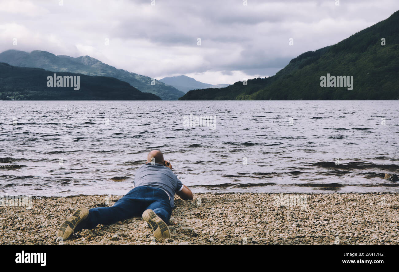 Travel photographer lying on pebble beach photographing Loch Lomond, Trossachs National Park, Stirlingshire/Dumbartonshire, Scotland Stock Photo