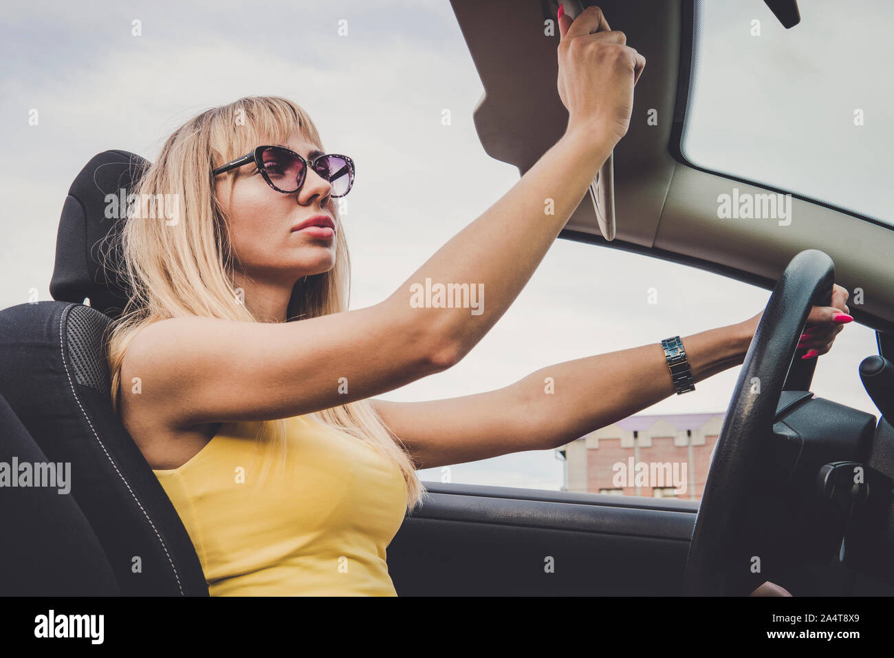 a woman admires her beauty in the reflection. Young cheerful woman driving car. Blonde girl looks in the mirror and preens. woman lowers the sun visor Stock Photo