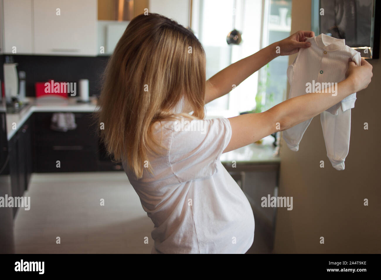 Pregnant happy woman examines small baby clothes with cheerful buttons, holds on outstretched arms. Pretending to try on. Homely atmosphere. Stock Photo