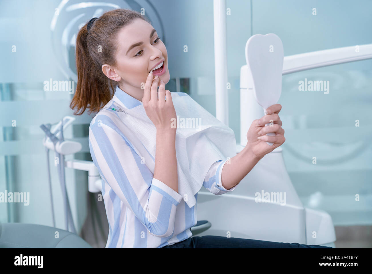 Happy woman satisfied with professional private clinic of dentistry. Charming and adorable woman looking at mirror with opened mouth, smiling. Client holding tooth shaped mirror. Stock Photo