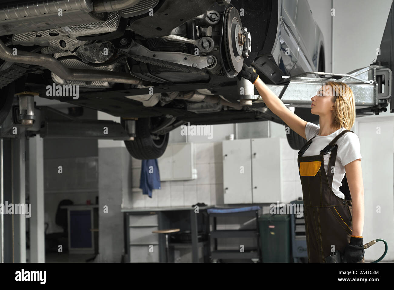 Beautiful young woman standing near lifted vehicle, observing and fixing brake discs with special tool. Girl wearing in white t shirt and coveralls, working in autoservice station. Stock Photo