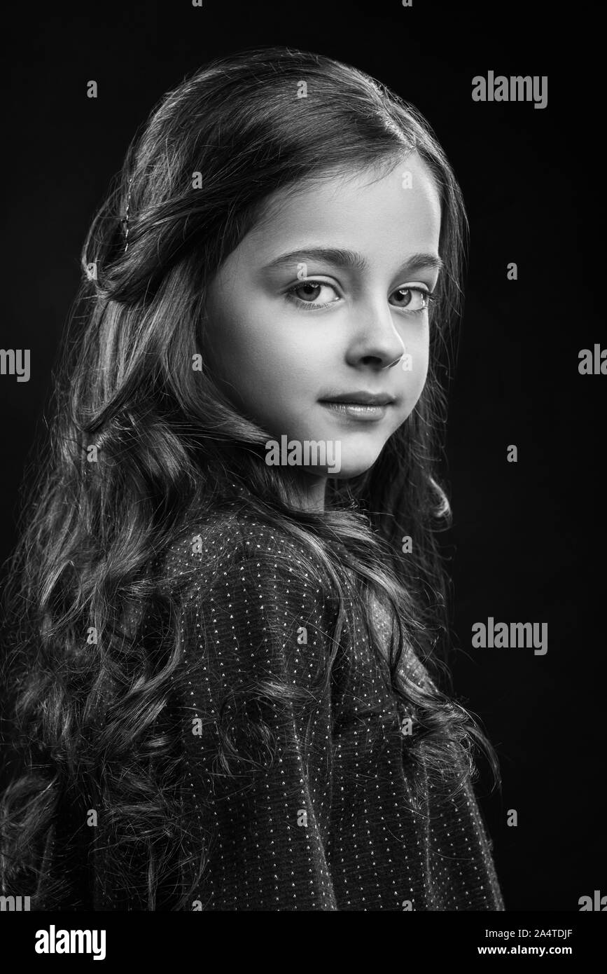 Portrait of charming long haired girl in dress posing on black isolated background. Pretty female child looking at camera and smiling in studio. Concept of beauty and elegance. Stock Photo