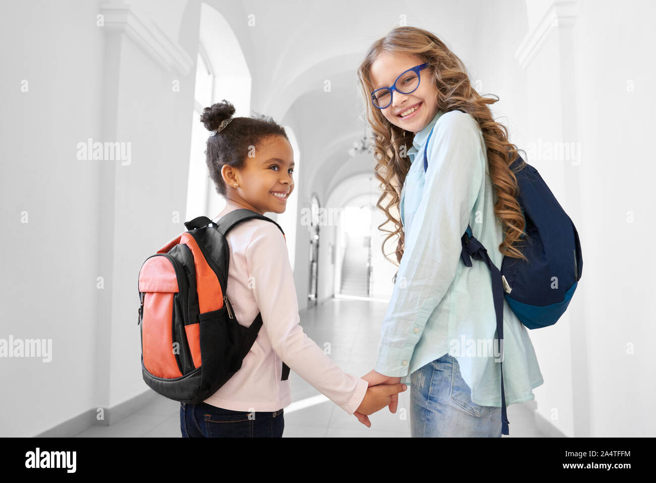 Two pretty school girls blonde Caucasian and brunette African holding each other by hands and smiling, going to lesson. Funny girls friends with deference in age spending time together. Stock Photo