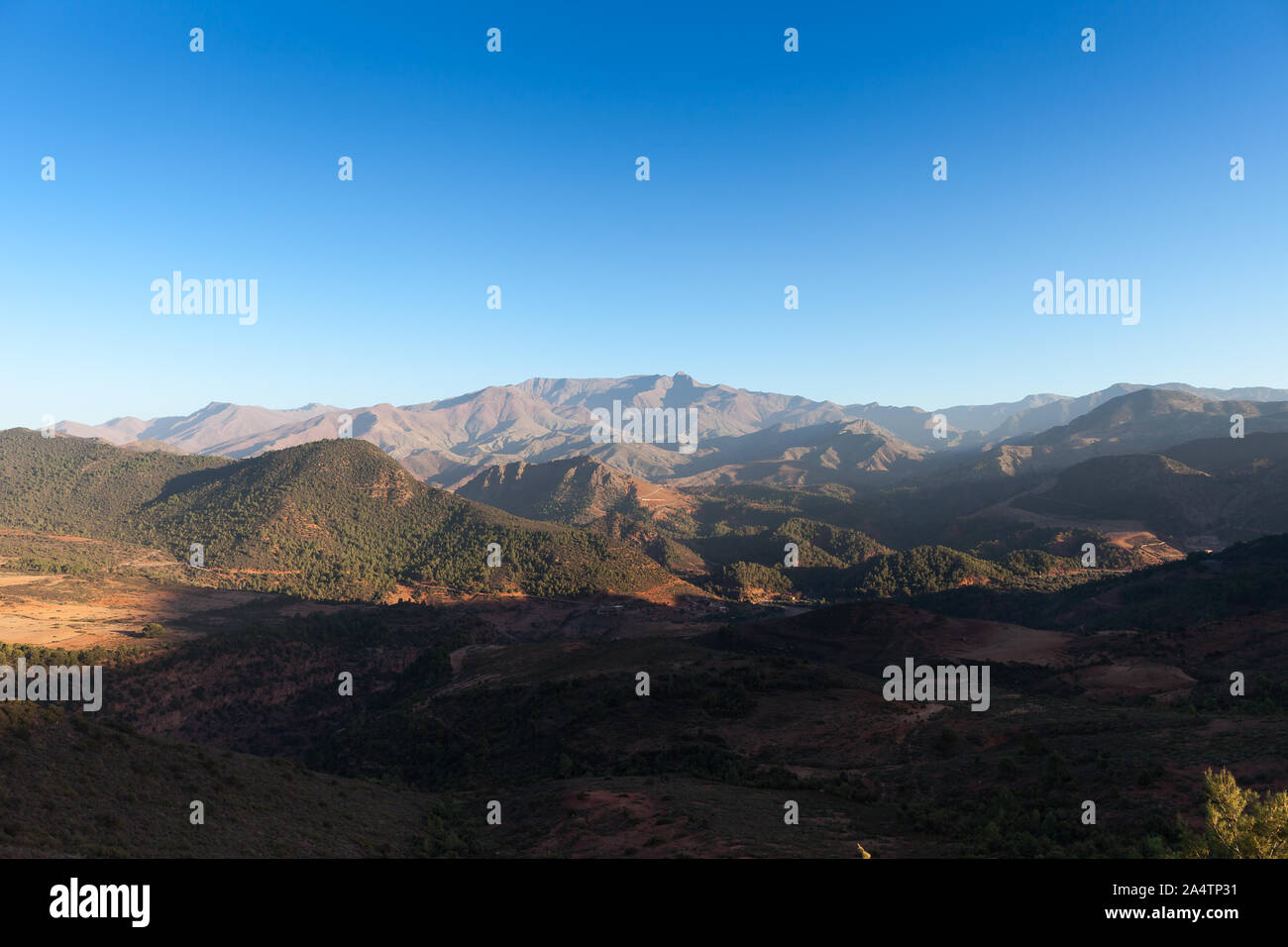 Panoramic viw of the High Atlas in Morocco with some green trees and a clear blue sky just before the sun is setting behind the mountains. Stock Photo