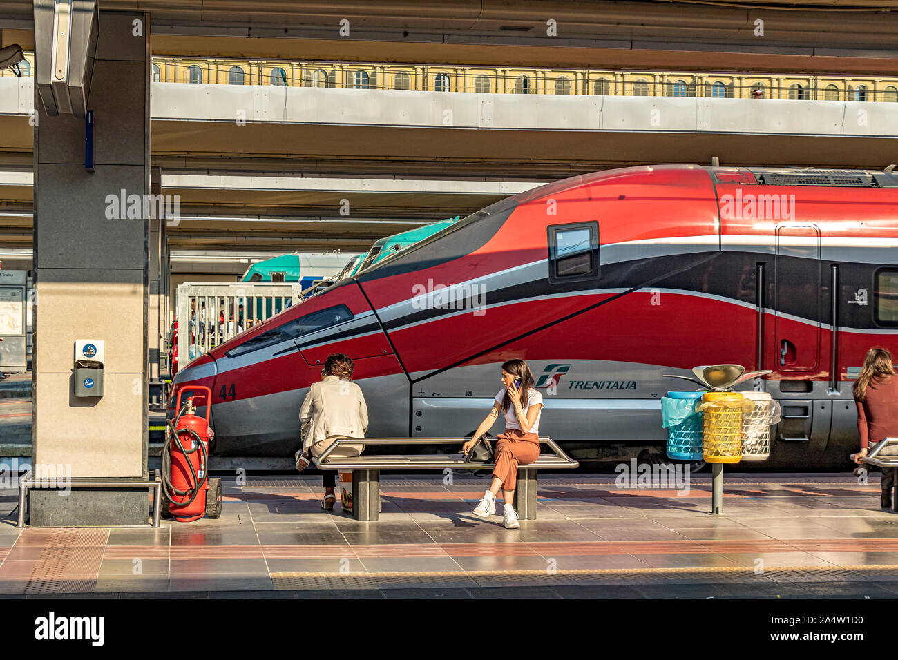 A girl sitting on a bench talking on her mobile phone in front of a Trenitalia Frecciarossa 1000 High Speed train at Porta Nuova railway station,Turin Stock Photo