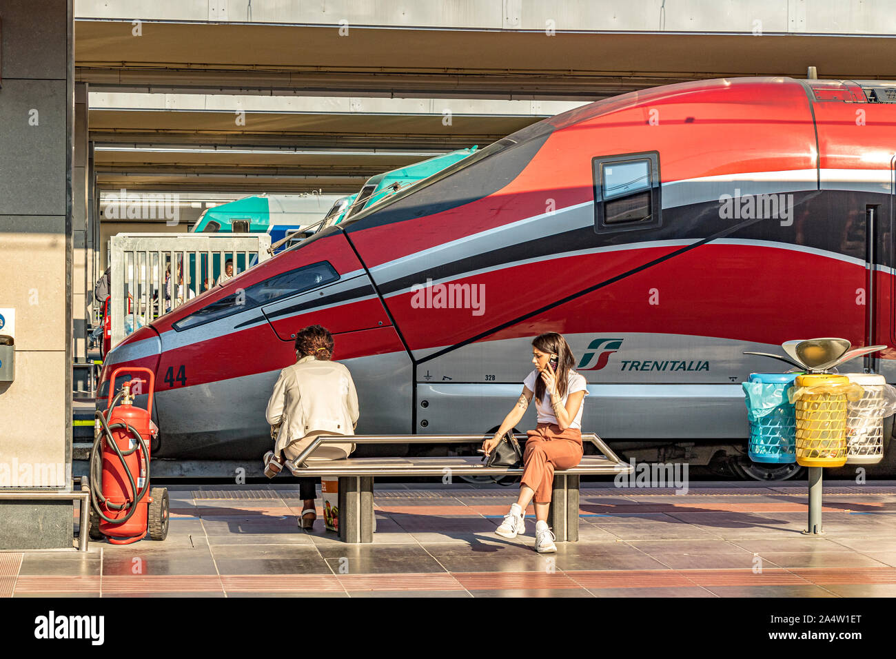 A girl sitting on a bench talking on her mobile phone in front of a Trenitalia Frecciarossa 1000 High Speed train at Porta Nuova railway station,Turin Stock Photo