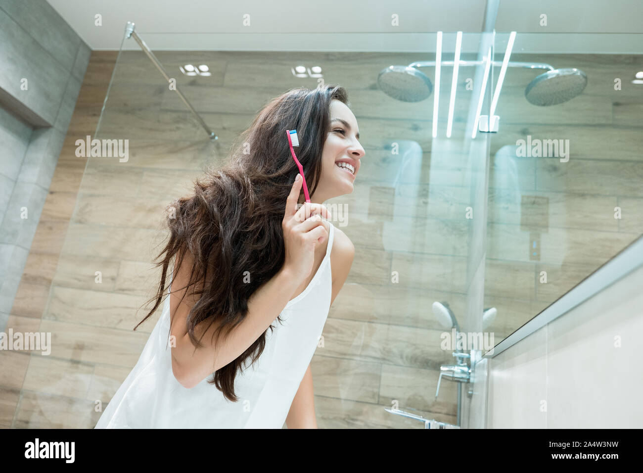 young beautiful woman standing in front of bathroom mirror while brushing her teeth Stock Photo
