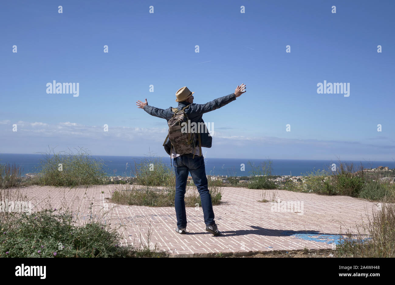 Male traveler with a backpack raised his arms high while standing on top of a mountain, against a blue sky. Stock Photo