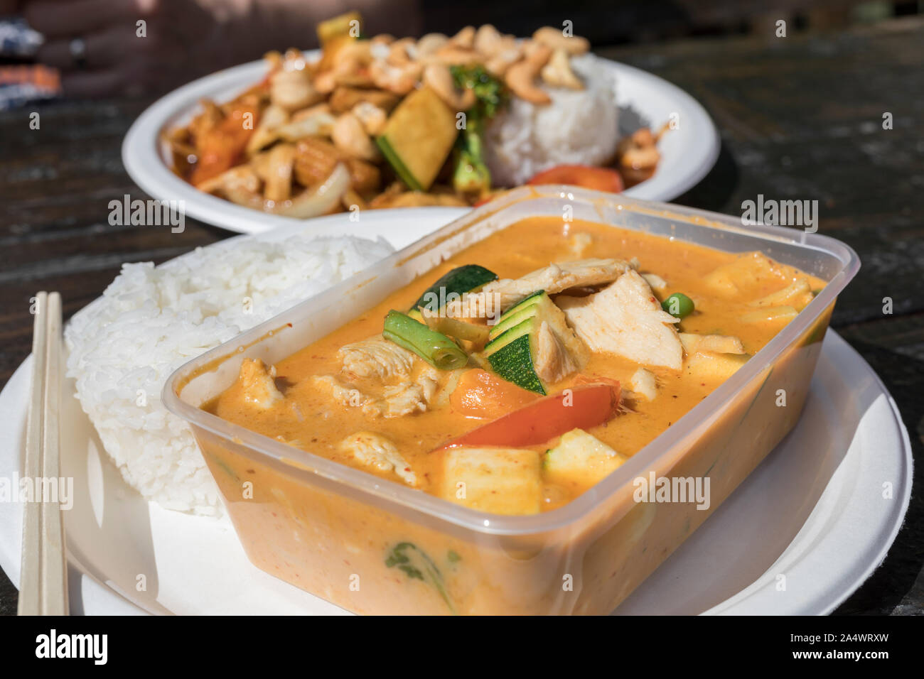 Thai take away food. One dish is in a plastic container and one is ready to eat on a disposable plate in the background. There are some wooden chopsti Stock Photo