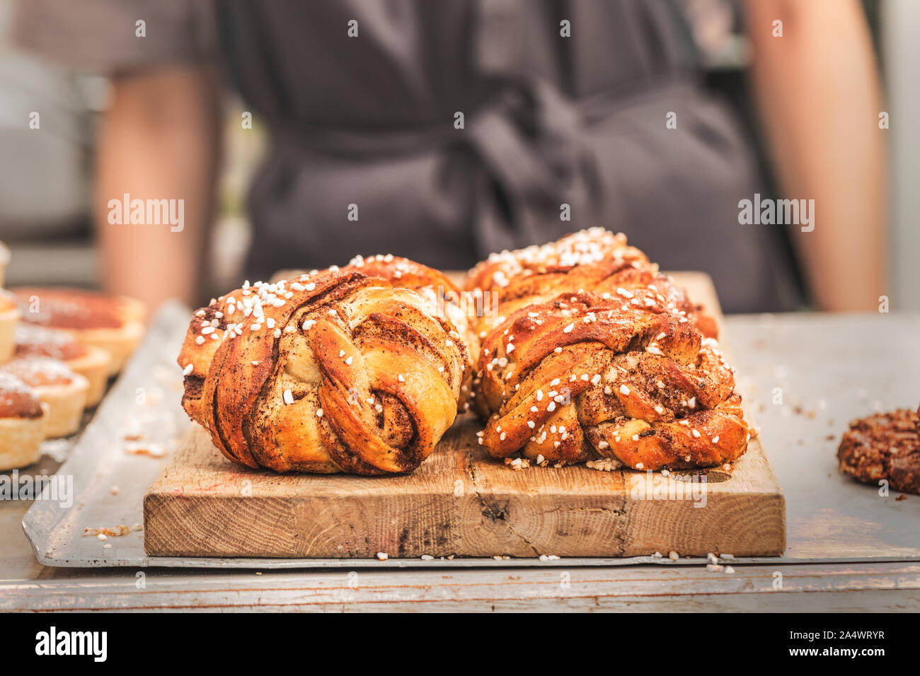 Twisted traditional Swedish cinnamon buns at a café. The sweet buns are on a wooden chopping board, and there is an  unrecognizable person with an apr Stock Photo