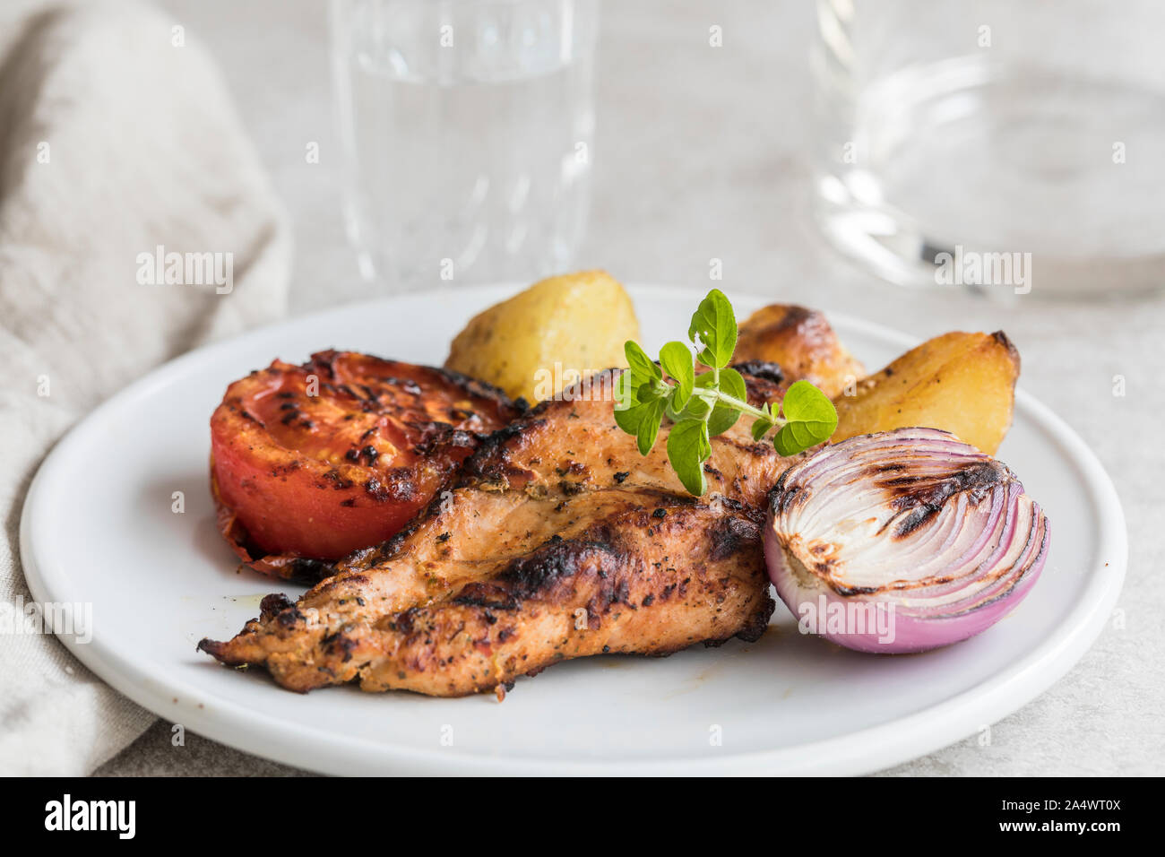 A fresh healthy meal with grilled chicken breast, roasted new potatoes, grilled tomato and grilled onion. The food is on a white plate with a glass of Stock Photo