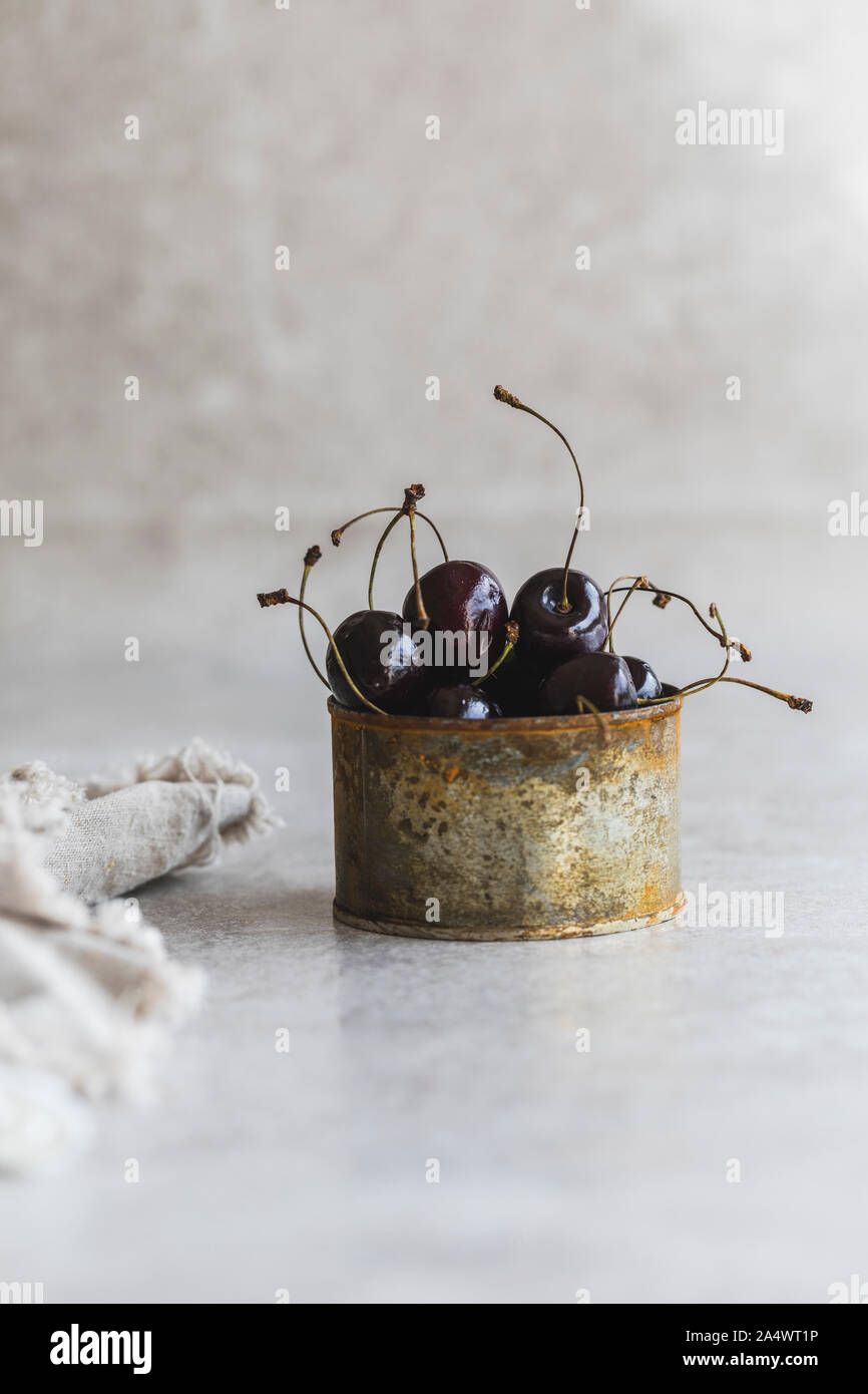 Black cherries with plant stems, in a old rusty tin.  This is on a gray background with a linen cloth next to the tin. Stock Photo